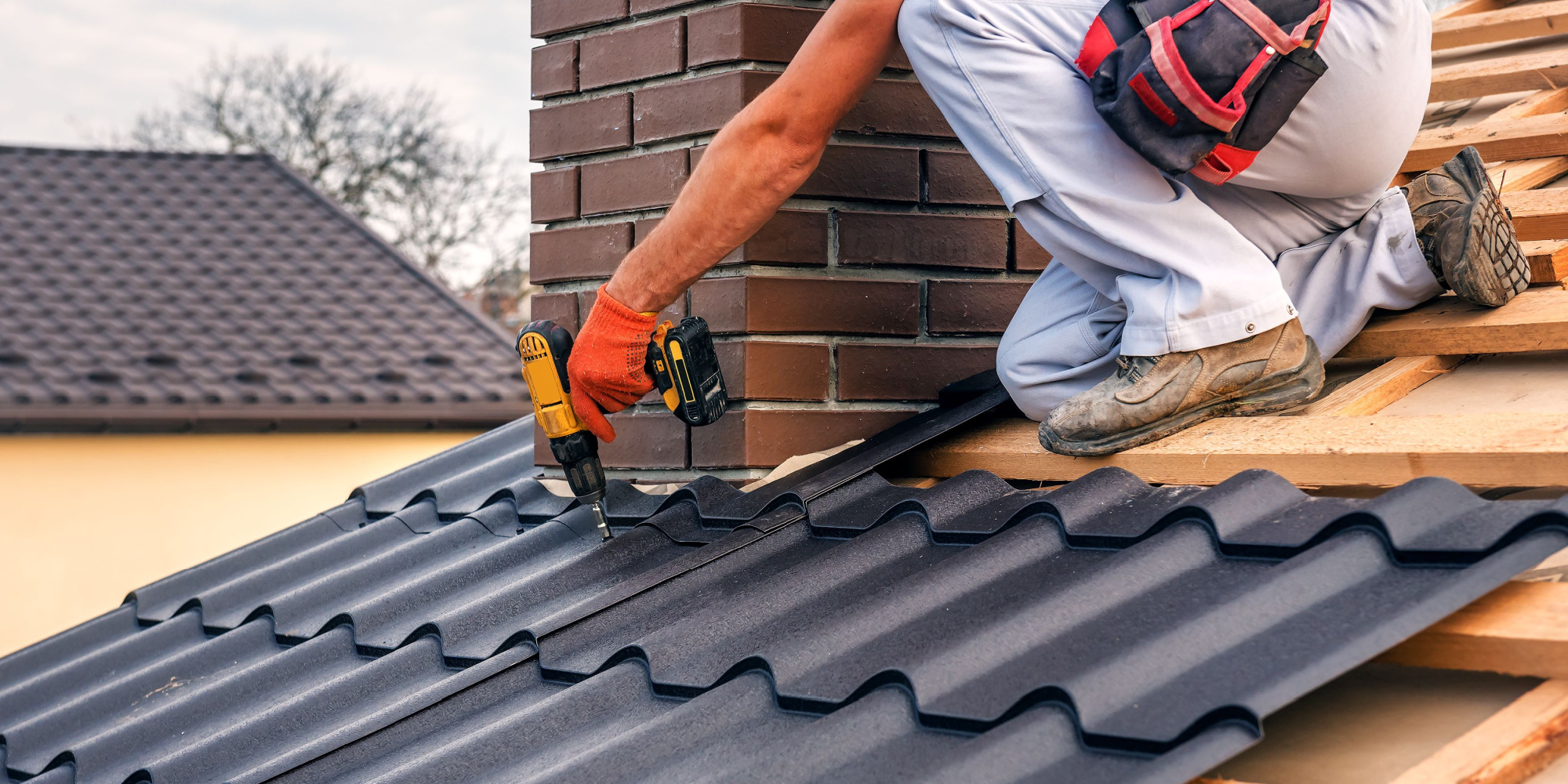 A roofer installs metal roofing panels on wood decking around a chimney.